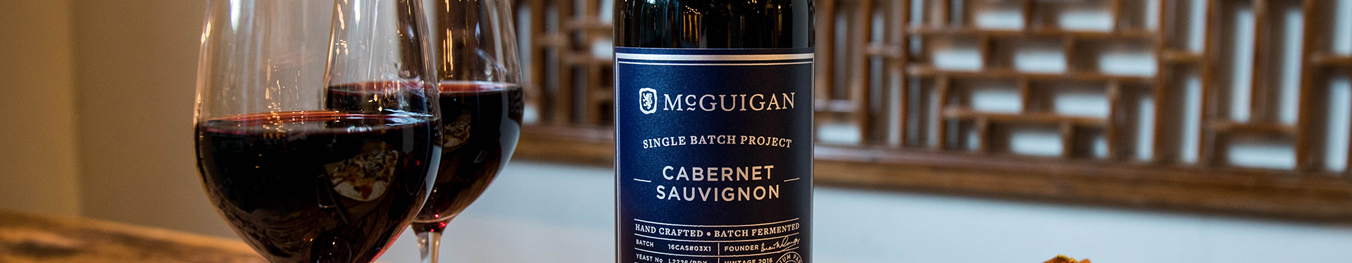 A bottle of McGuigan Single Project Cabernet Sauvignon and with two glasses of wine Black Label Red Mid in a dinner table setting.